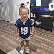 Juelz joined the competition — help win amazing prizes! toddler, child, indoor, flooring, door, cabinet, tv, sports_jersey, white_shorts, white_sneakers, hair_bun, wood_floor, living_room, person, furniture, casual_clothing, young_child, standing, expression, home