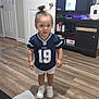 toddler, child, indoor, flooring, door, cabinet, tv, sports_jersey, white_shorts, white_sneakers, hair_bun, wood_floor, living_room, person, furniture, casual_clothing, young_child, standing, expression, home