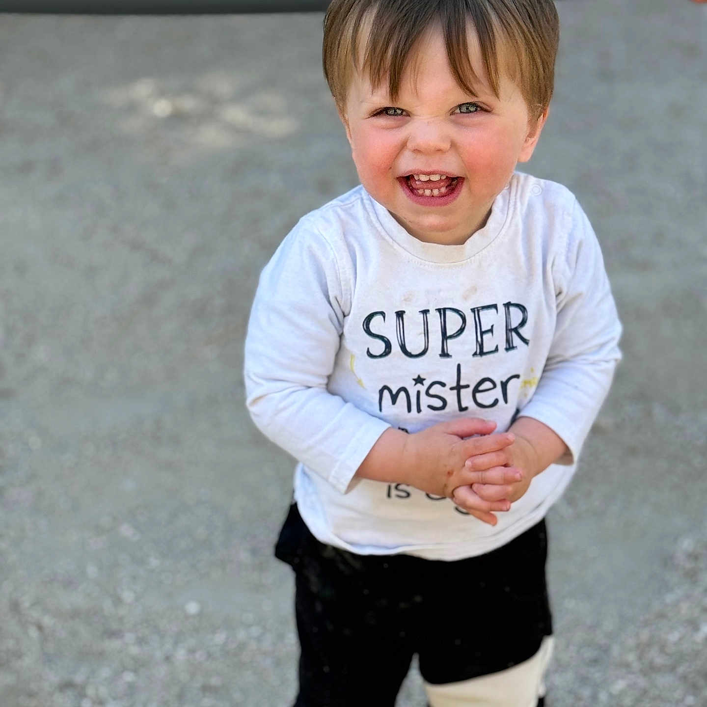 Ayden participe au concours pour gagner de l'argent avec cette photo : black_pants, casual, child, clothing, cute, daylight, face, gravel, hands_clasped, happy, outdoor, person, playground, portrait, short_hair, smiling, standing, toddler, white_shirt, young