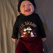 Brixton is registered to the contest to win money with this photo: baby, smiling, laughing, black_beanie, shirt, graphic_tee, lil_shark, pants, maroon_pants, lying_down, bed, blanket, happy, child, infant, cute, tongue_out, face, person, clothing