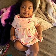Nevaeh joined the competition — help win amazing prizes! baby, infant, pink_dress, cherry_pattern, blanket, couch, fluffy_slippers, pacifier, phone_case, curly_hair, smiling, lying_down, indoor, soft_texture, child, person, cute, cozy, portrait, happy