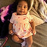 baby, infant, pink_dress, cherry_pattern, blanket, couch, fluffy_slippers, pacifier, phone_case, curly_hair, smiling, lying_down, indoor, soft_texture, child, person, cute, cozy, portrait, happy