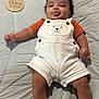 baby, infant, child, cute, tongue_out, white_overalls, orange_shirt, quilted_bedspread, wooden_plaque, three_months, smiling, lying_down, portrait, indoors, soft_lighting, playful, face, hands, legs, clothing