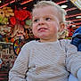 Rafaël a rejoint le concours — aidez-le/la à gagner de superbes lots ! toddler, child, face, smile, striped_shirt, hand, carnival, stuffed_animals, toy, colorful, neon_lights, indoor, person, clothing, playful, amusement_park, game_booth, curly_hair, holding, funfair