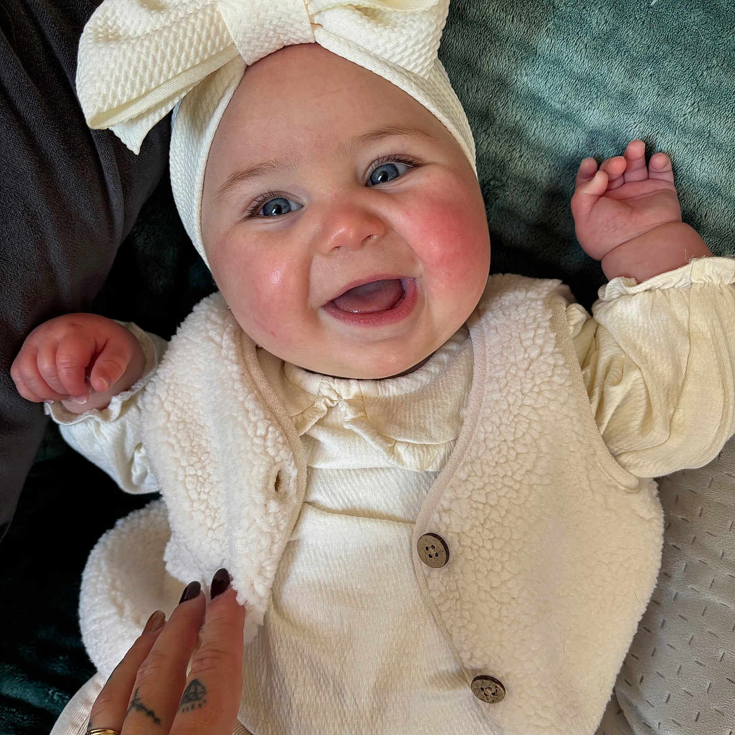 Adrya participe au concours pour gagner de l'argent avec cette photo : adult_hand, baby, blanket, blue_eyes, bow, buttons, closeup, fabric_texture, hand, happy, headband, indoor, infant, onesie, portrait, ring, rosy_cheeks, smile, tattoo, vest