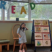 Dawn is registered to the contest to win money with this photo: animal_illustrations, carpet, chair, child, classroom, display_board, dress, educational, girl, indoor, letters, library, nature_outside, pink_shoes, playful, poster, reading_area, smiling, window, wooden_furniture