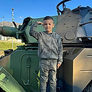 Leonardo participe au concours pour gagner de l'argent avec cette photo : child, boy, salute, military_vehicle, outdoor, daylight, blue_sky, mountains, gray_tracksuit, sneakers, standing, portrait, young, casual_clothing, vehicle, green, metal, serious, person, urban