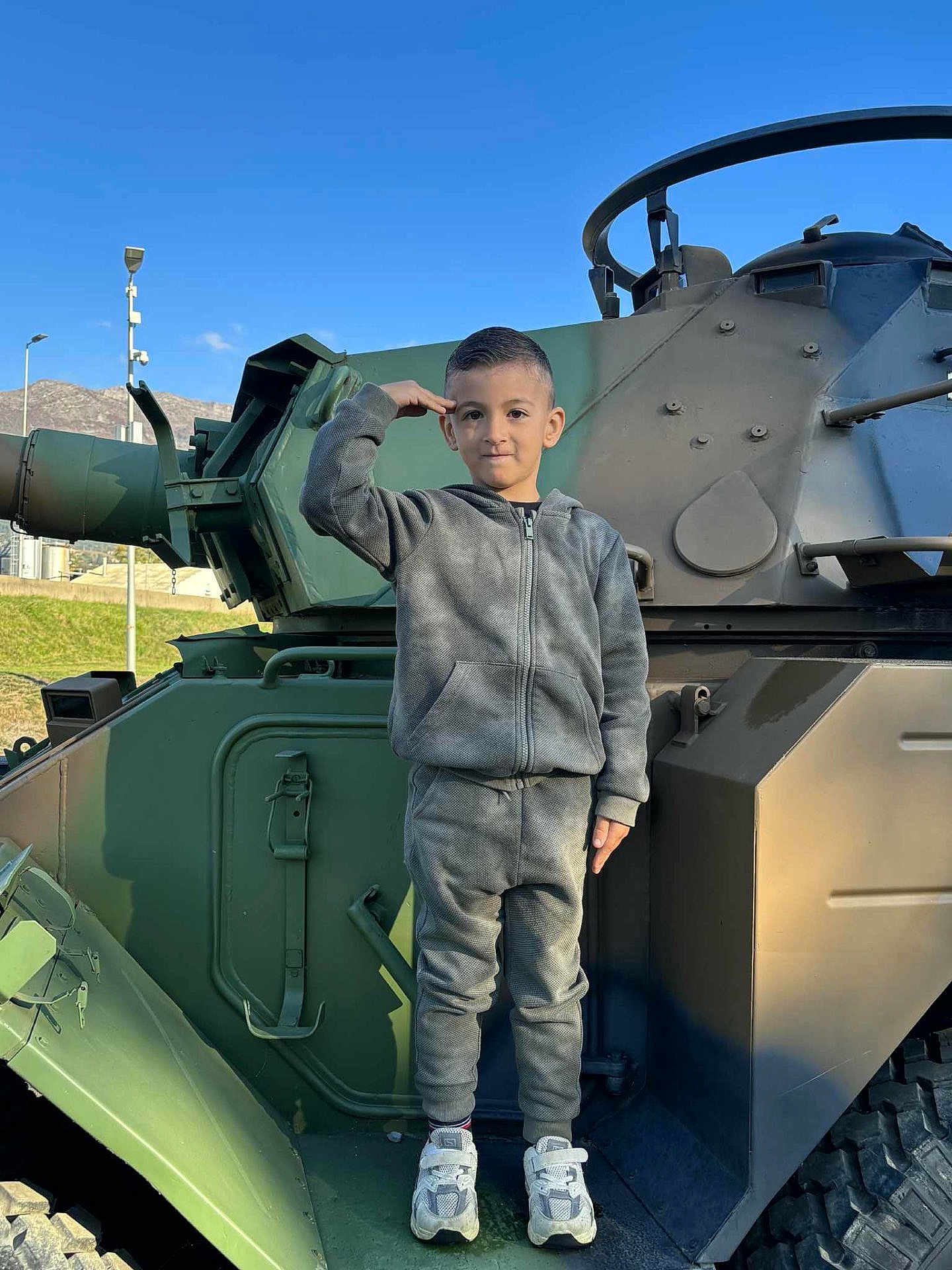 Leonardo participe au concours pour gagner de l'argent avec cette photo : child, boy, salute, military_vehicle, outdoor, daylight, blue_sky, mountains, gray_tracksuit, sneakers, standing, portrait, young, casual_clothing, vehicle, green, metal, serious, person, urban