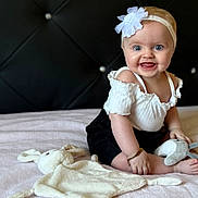 Ariana joined the competition — help win amazing prizes! baby, bed, black_shorts, blue_eyes, child, cute, happy, headband, indoors, infant, playful, plush_toy, portrait, sitting, smiling, soft_blanket, tufted_headboard, white_flower, white_top, young_child