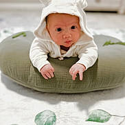 Jaxson J is registered to the contest to win money with this photo: baby, infant, hoodie, white_clothing, cushion, green_cushion, lying_down, face, hands, curious, indoors, carpet, blurred_background, cute, child, soft_lighting, baby_boy, baby_girl, hood_with_ears, person
