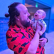 Wren is registered to the contest to win money with this photo: baby, child, infant, adult, father, parent, family, portrait, smiling, laughing, holding, cuddling, beard, long_hair, shirt, patterned_clothing, indoor, colorful_lighting, blue_lighting, happy