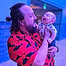 baby, child, infant, adult, father, parent, family, portrait, smiling, laughing, holding, cuddling, beard, long_hair, shirt, patterned_clothing, indoor, colorful_lighting, blue_lighting, happy