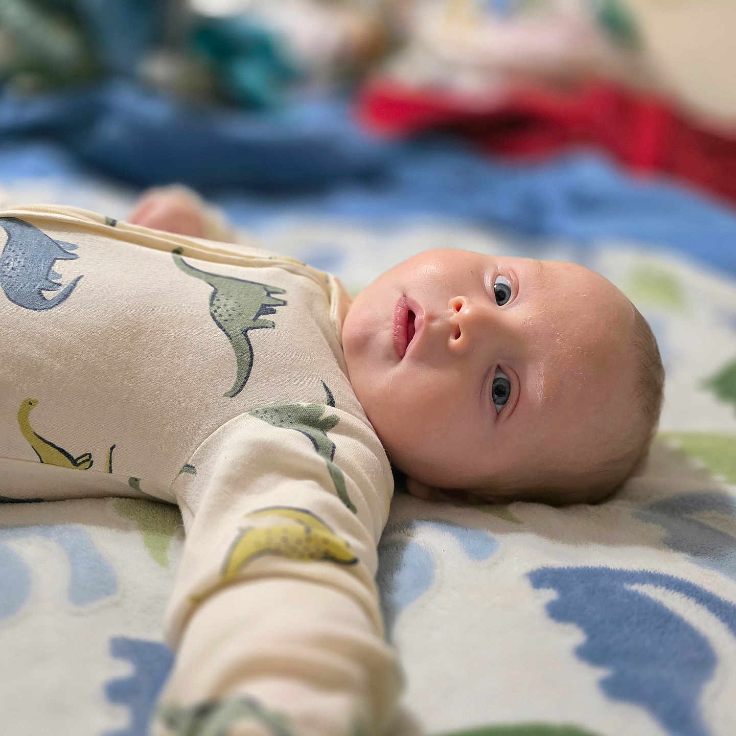 Wren joined the competition — help win amazing prizes! baby, infant, child, portrait, lying_down, bed, blanket, dinosaur_pattern, onesie, clothing, face, eyes, open_mouth, smiling, indoor, bedroom, cozy, colorful, close_up, photography