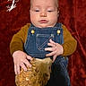 Wren is registered to the contest to win money with this photo: baby, infant, child, portrait, lying_down, red_background, denim_overalls, brown_shirt, feathers, golden_prop, costume, serious_expression, face, hands, blue_eyes, indoor, studio_portrait, close_up, decorative_design, photography