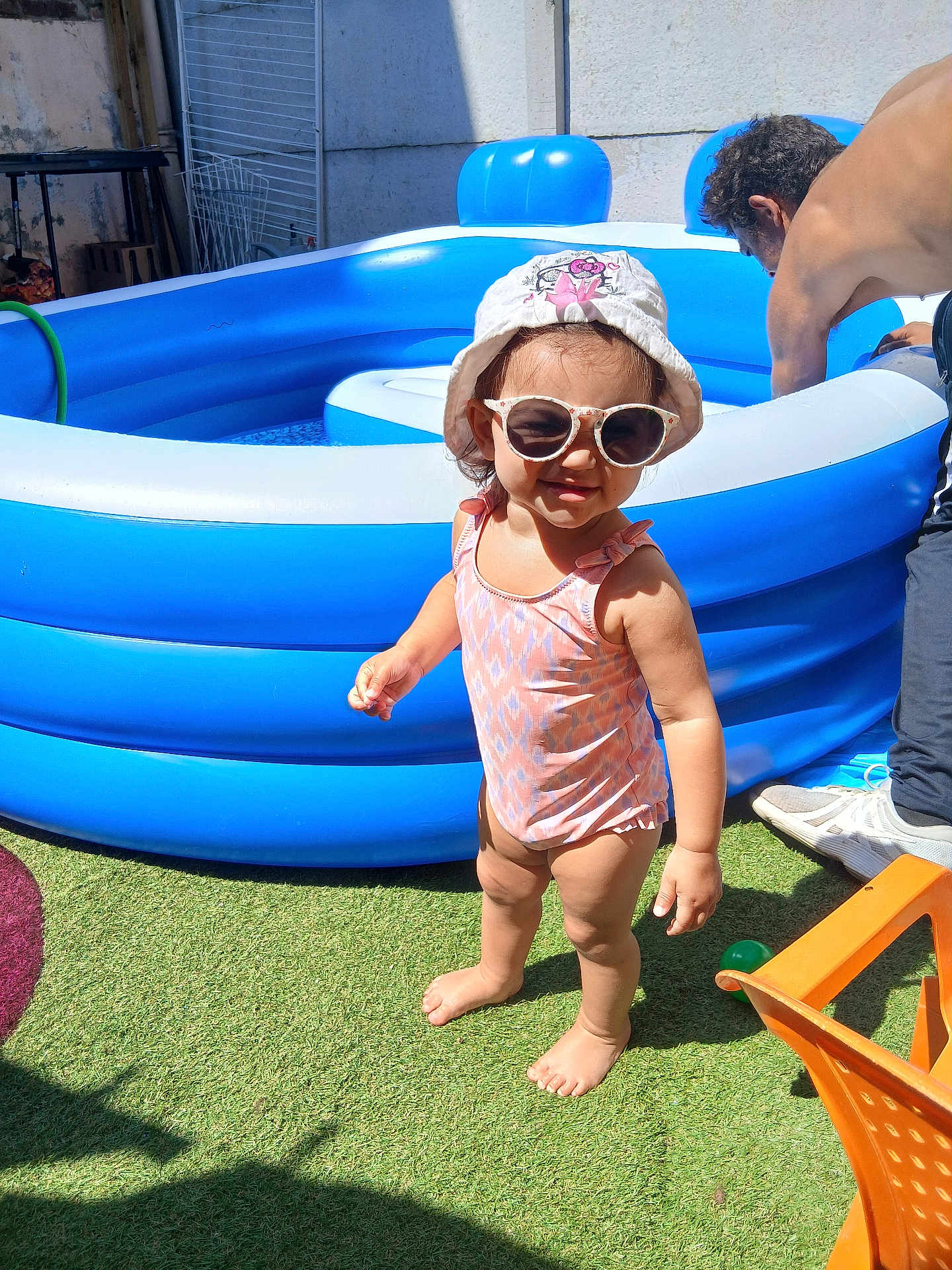 Inaya participe au concours pour gagner de l'argent avec cette photo : toddler, child, swimsuit, sunglasses, hat, barefoot, grass, inflatable_pool, summer, sunlight, outdoor, man, adult, backyard, water, toy, blue, pink, shadow, play
