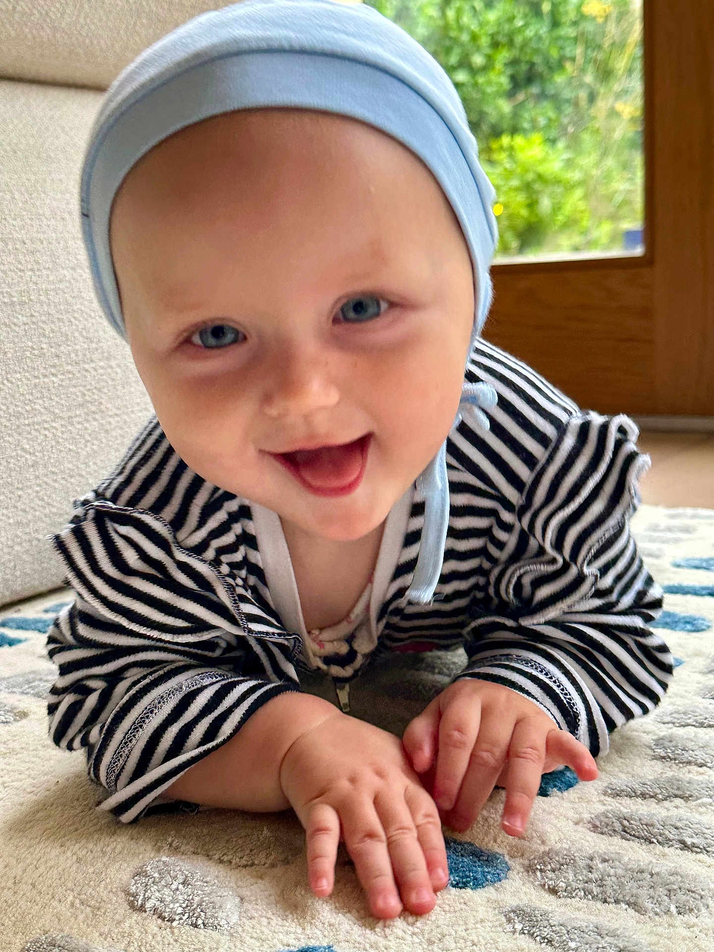 Lily is registered to the contest to win money with this photo: baby, infant, crawling, smiling, striped_clothing, blue_hat, carpet, indoor, face, hands, cute, child, happy, person, floor, home, playing, bright_eyes, closeup, natural_light