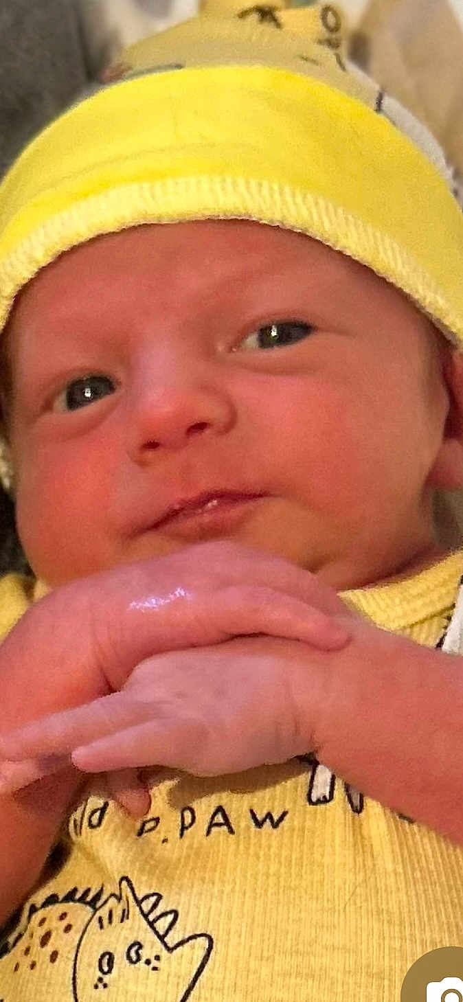 Braxton is registered to the contest to win money with this photo: baby, newborn, infant, yellow_hat, yellow_clothing, dinosaur_print, hands, close_up, face, cute, soft_skin, curious_expression, indoor, child, person, portrait, young, sleepy_eyes, resting, babywear