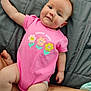baby, infant, pink_onesie, flower_graphic, text, gray_quilt, arm, skin, cute, chubby_cheeks, laying_down, indoor, child, person, apparel, soft_lighting, casual, closeup, happy, young
