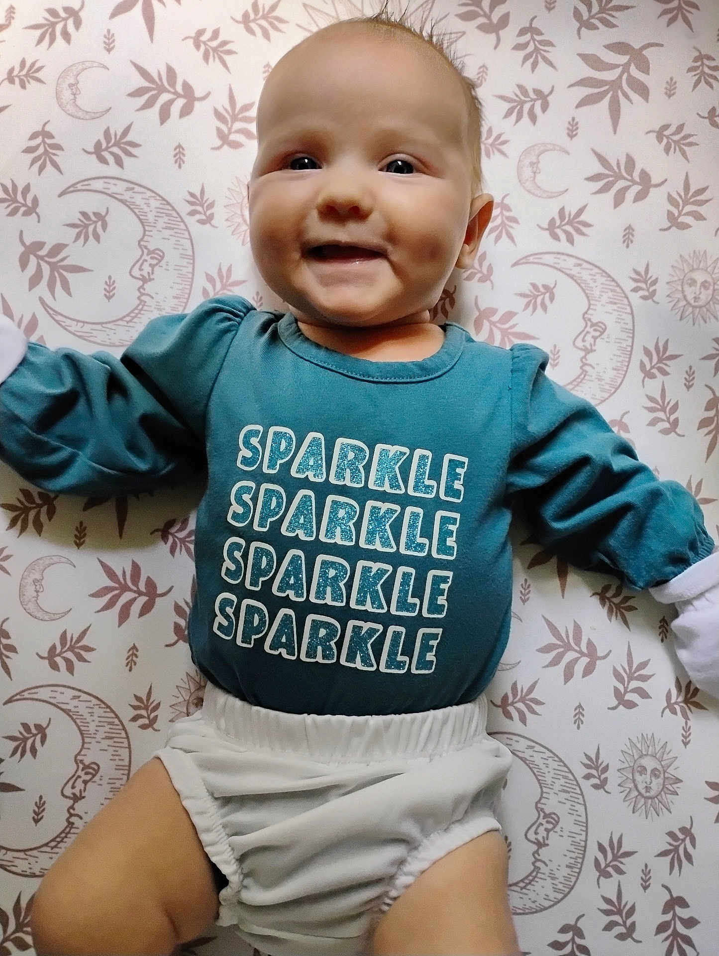 Laynie joined the competition — help win amazing prizes! baby, child, smiling, blue_shirt, text_on_clothing, patterned_blanket, moon_pattern, leaf_pattern, infant, happy, lying_down, cute, face, white_diaper, long_sleeves, indoor, soft_lighting, portrait, person, young_child