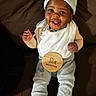 baby, bib, brown_couch, celebration, child, couch, cozy_clothing, cute, feet, happy, hat, indoor, infant, milestone, person, portrait, sitting, smiling, socks, wooden_sign