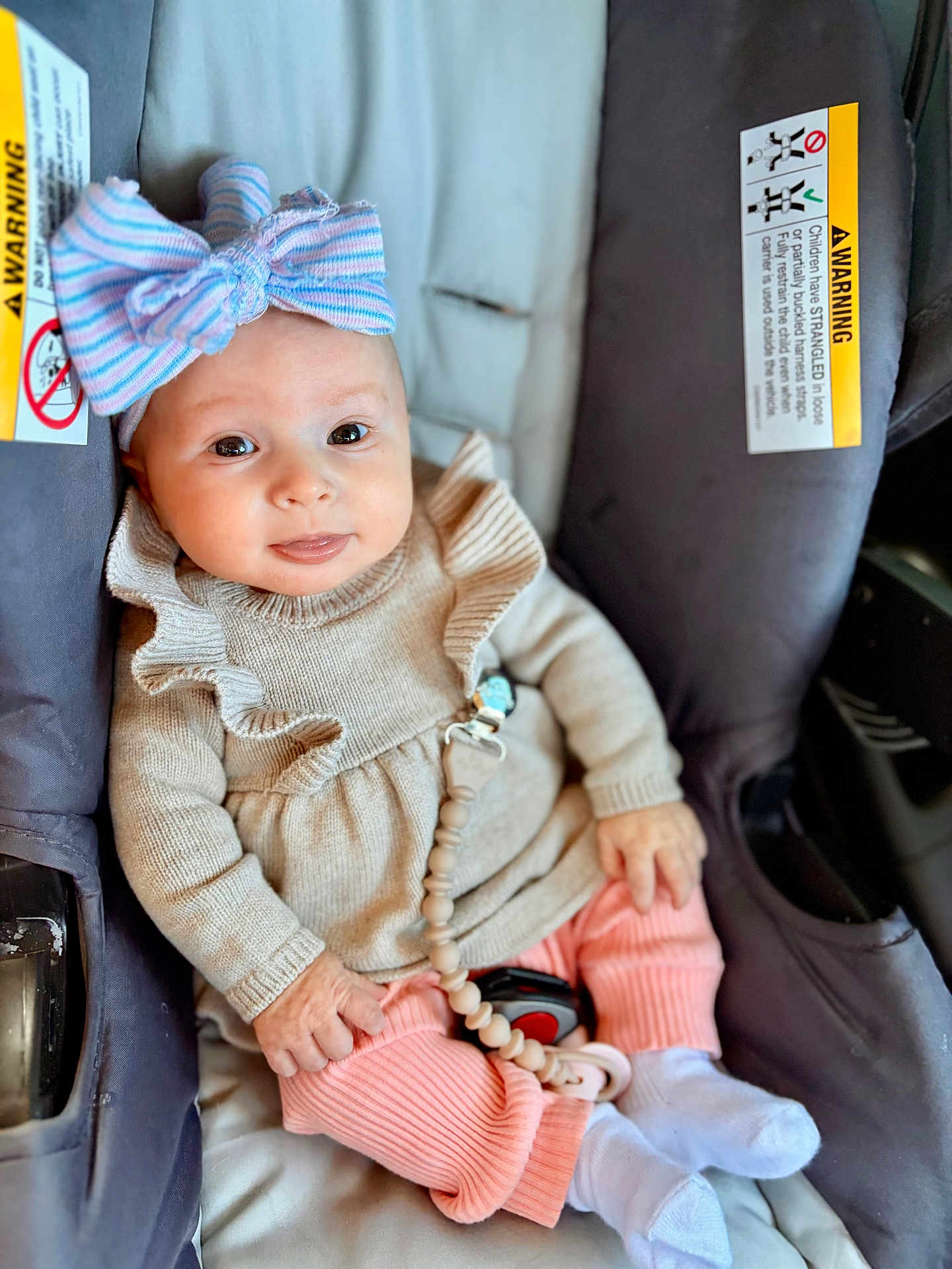 Viviana is registered to the contest to win money with this photo: baby, child, car_seat, bow, smile, pink_pants, beige_sweater, white_socks, infant, cute, happy, seated, indoors, clothing, face, hand, toy, accessory, portrait, person