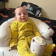 Luca-Lee joined the competition — help win amazing prizes! baby, infant, yellow_onesie, smiling, inflatable_chair, cup_lid, bedroom, bed, blanket, pillow, patterned_fabric, cozy, child, indoor, person, cute, happy, sitting, footed_pajamas, toy