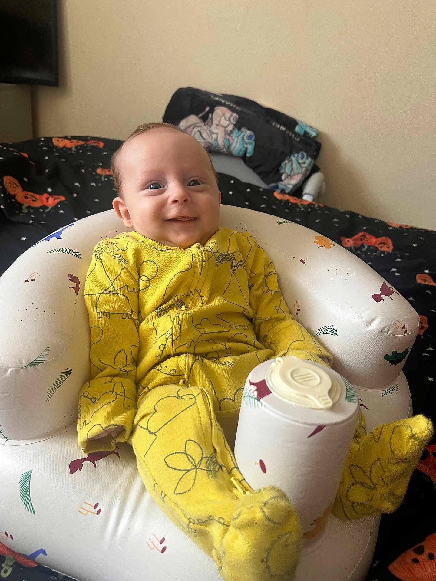 Luca-Lee joined the competition — help win amazing prizes! baby, infant, yellow_onesie, smiling, inflatable_chair, cup_lid, bedroom, bed, blanket, pillow, patterned_fabric, cozy, child, indoor, person, cute, happy, sitting, footed_pajamas, toy