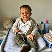 Taho a rejoint le concours — aidez-le/la à gagner de superbes lots ! toddler, child, smiling, sitting, white_towel, changing_table, knit_cardigan, polo_shirt, olive_pants, sneakers, pacifier, wooden_beads, baby_care_products, folded_clothes, indoor, happy, baby_accessories, wet_hair, person, cute