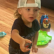 Jaden is registered to the contest to win money with this photo: child, toddler, cap, backwards_cap, curly_hair, toy_truck, green_toy, wooden_floor, indoor, couch, black_tshirt, blue_jeans, reaching_hand, playing, floor, expression, person, casual_clothing, young_child, headwear