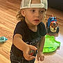 child, toddler, cap, backwards_cap, curly_hair, toy_truck, green_toy, wooden_floor, indoor, couch, black_tshirt, blue_jeans, reaching_hand, playing, floor, expression, person, casual_clothing, young_child, headwear