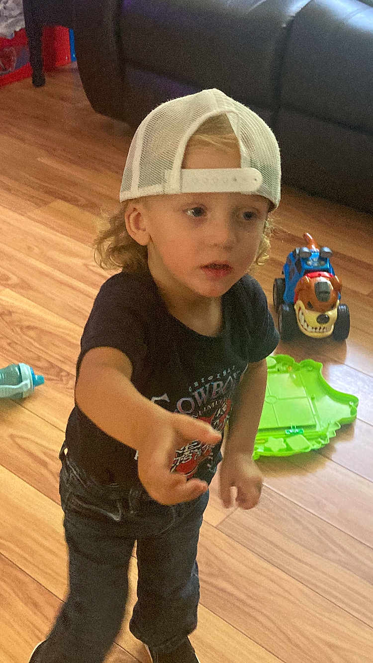 Jaden is registered to the contest to win money with this photo: child, toddler, cap, backwards_cap, curly_hair, toy_truck, green_toy, wooden_floor, indoor, couch, black_tshirt, blue_jeans, reaching_hand, playing, floor, expression, person, casual_clothing, young_child, headwear