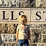 blond_hair, casual_clothing, child, concrete, daylight, engraved_text, jeans, outdoor, person, portrait, shadow, sidewalk, smiling, sneakers, standing, stone_wall, sunlight, toddler, tshirt, young_child