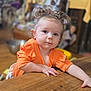 Brooklyn is registered to the contest to win money with this photo: baby, child, curly_hair, cute, expression, face, furniture, hairstyle, hand, home, indoor, leaning, orange_clothing, person, portrait, skin, table, toddler, wood, young_child