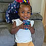 toddler, child, smiling, indoor, sofa, cup, backpack, hair_beads, white_shirt, yellow_pants, carpet, earrings, furniture, home, happy, person, portrait, young_child, seated, casual_clothing