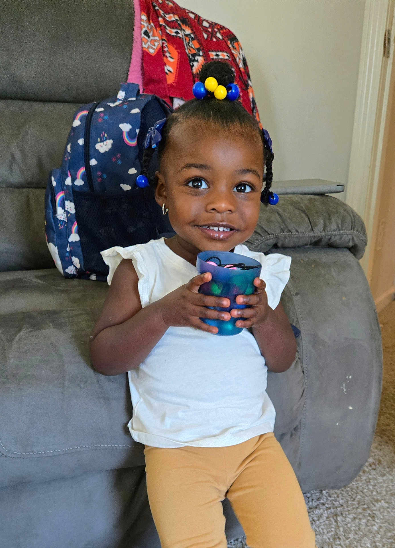 Jannah joined the competition — help win amazing prizes! toddler, child, smiling, indoor, sofa, cup, backpack, hair_beads, white_shirt, yellow_pants, carpet, earrings, furniture, home, happy, person, portrait, young_child, seated, casual_clothing