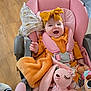 baby, child, smiling, stroller, pink, yellow, headband, bow, blanket, plush_toy, toy, happy, indoor, clothing, seat_belt, wood_floor, person, cute, infant, comfort