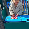 bench, blue_panel, candid, child, coat, face, hands, hood, jacket, outdoor, park, play_equipment, playground, railing, smile, sunlit, toddler, toy, trees, woodchips