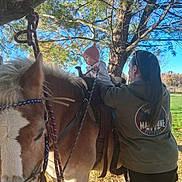 Whitlee is registered to the contest to win money with this photo: baby, child, adult, pony, horse, horseback_riding, outdoor, park, pine_tree, saddle, bridle, reins, winter_clothing, pink_hat, animal, riding, family, nature, sunlight, blue_sky