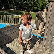 Mylo a rejoint le concours — aidez-le/la à gagner de superbes lots ! beige_pants, casual_clothing, child, curious, daylight, greenery, nike_shirt, outdoor, park, person, play, playground, shadow, standing, sunny, toddler, trees, wheel, white_shoes, wooden_structure