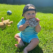Bryce is registered to the contest to win money with this photo: baby, child, grass, outdoor, sunlight, cap, blue_clothing, plaque, toy, teddy_bear, milestone, chewing, candid, summer, sitting, barefoot, portrait, infant, nature, playtime