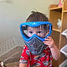 toddler, child, mask, goggles, blue_mask, red_shirt, racing_print, indoor, toy_container, wooden_shelf, curious, face, hand, short_hair, person, play, young_child, casual_clothing, home, portrait