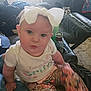 baby, bow, child, couch, floral_pants, indoor, living_room, person, shirt, sitting, smile, white_bow, wide_eyes, bottle, cup_holder, blanket, carpet, furniture, relaxed, casual
