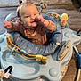 baby, child, activity_jumper, blue, indoor, flooring, wooden_floor, carpet, toy, smiling, thumb_in_mouth, winking, bracelet, clothing, person, playful, home, furniture, seat, cute