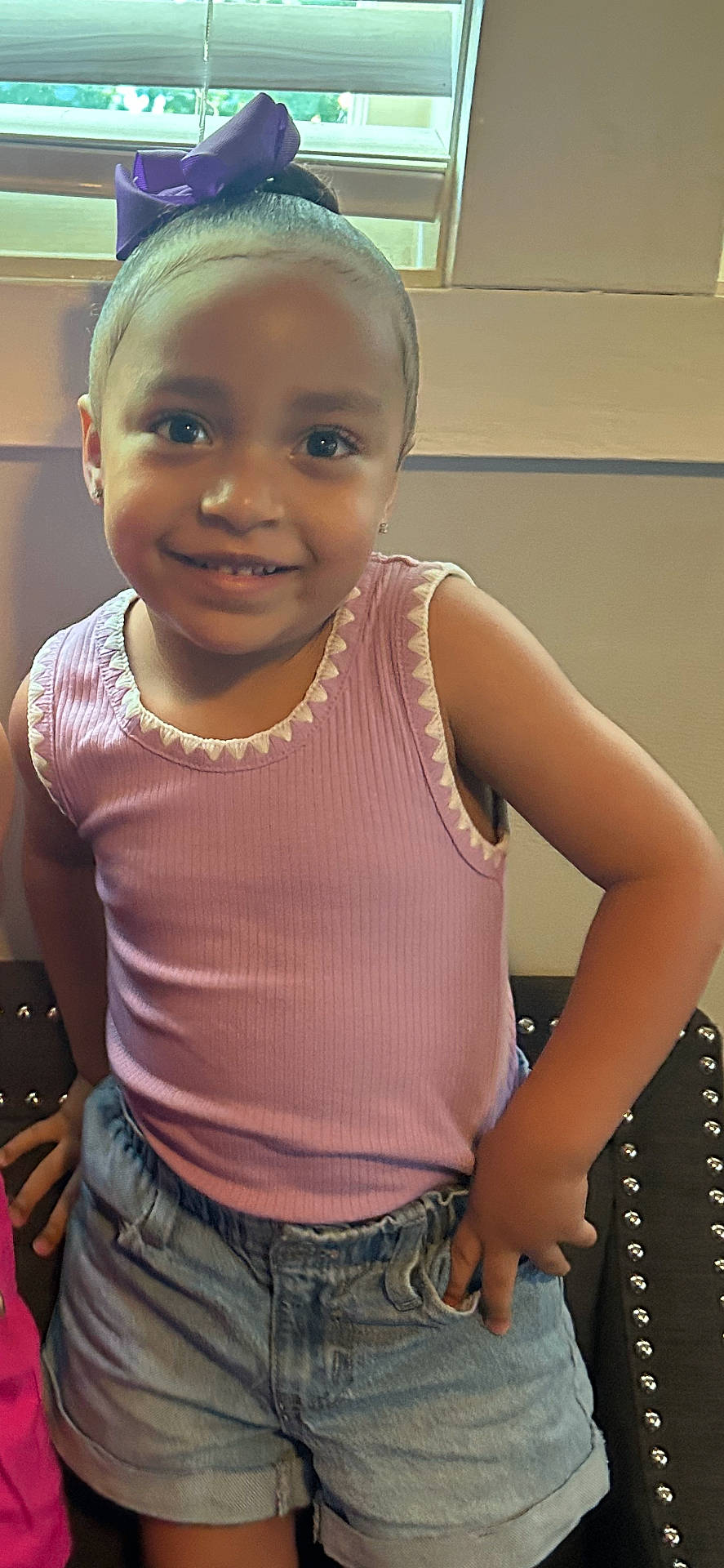 accessories, casual, child, childhood, clothing, cute, denim_shorts, fashion, girl, hand_on_hip, happy, indoor, person, pink_top, portrait, posing, purple_bow, smiling, standing, window