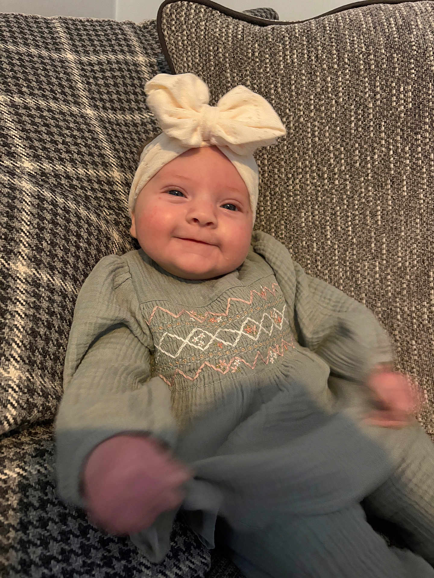 Willow is registered to the contest to win money with this photo: baby, child, smiling, headband, bow, dress, couch, textile, pattern, hand, face, indoor, happy, infant, apparel, seated, person, cute, portrait, motion_blur