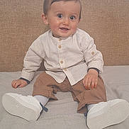 Ayoub participe au concours pour gagner de l'argent avec cette photo : baby, child, smiling, sitting, indoor, bed, white_shoes, beige_headboard, light_shirt, brown_pants, happy, portrait, person, cute, young_child, clothing, footwear, face, eyes, hands