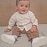 baby, child, smiling, sitting, indoor, bed, white_shoes, beige_headboard, light_shirt, brown_pants, happy, portrait, person, cute, young_child, clothing, footwear, face, eyes, hands