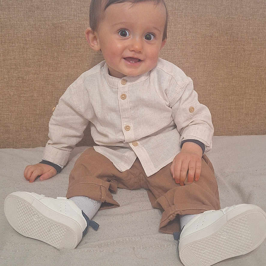 Ayoub participe au concours pour gagner de l'argent avec cette photo : baby, child, smiling, sitting, indoor, bed, white_shoes, beige_headboard, light_shirt, brown_pants, happy, portrait, person, cute, young_child, clothing, footwear, face, eyes, hands