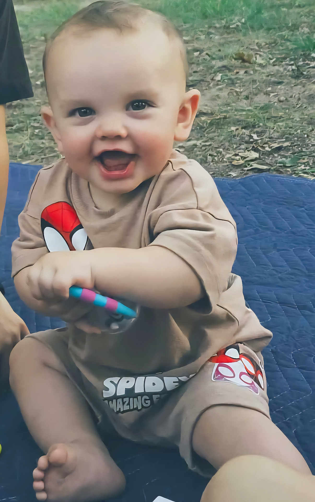 Theodore joined the competition — help win amazing prizes! baby, child, smiling, outdoor, grass, toy, clothing, spiderman, sitting, foot, hand, quilt, happy, cute, person, infant, face, head, playful, barefoot