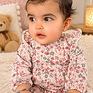Myra joined the competition — help win amazing prizes! baby, child, infant, portrait, sitting, floral_dress, pink_clothing, hair_bow, wide_eyes, looking_away, indoors, cozy, blanket, teddy_bear, flowers, lamp, soft_lighting, nursery, carpet, happy
